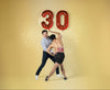 Two people embracing with red '30' balloons against a yellow background